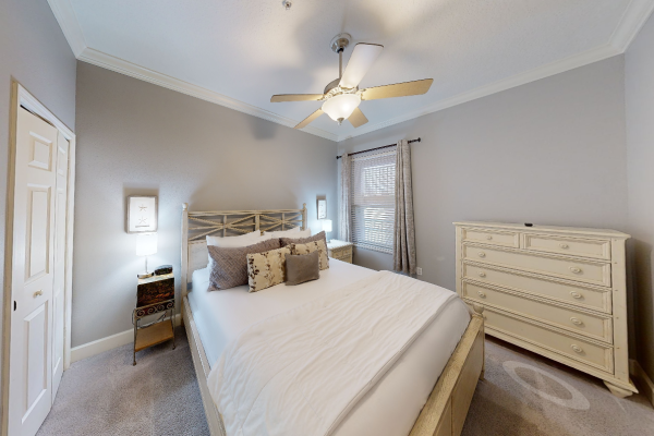A cozy bedroom with a large bed, decorative pillows, two side tables with lamps, a ceiling fan, and a wooden dresser near the window.
