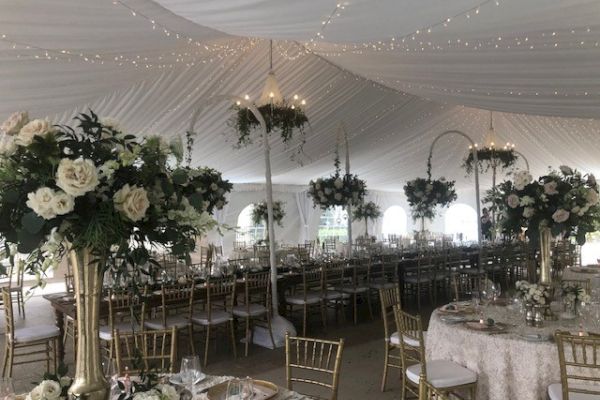 A beautifully decorated wedding tent with white drapes, floral arrangements, and gold chairs, creating an elegant and festive atmosphere.