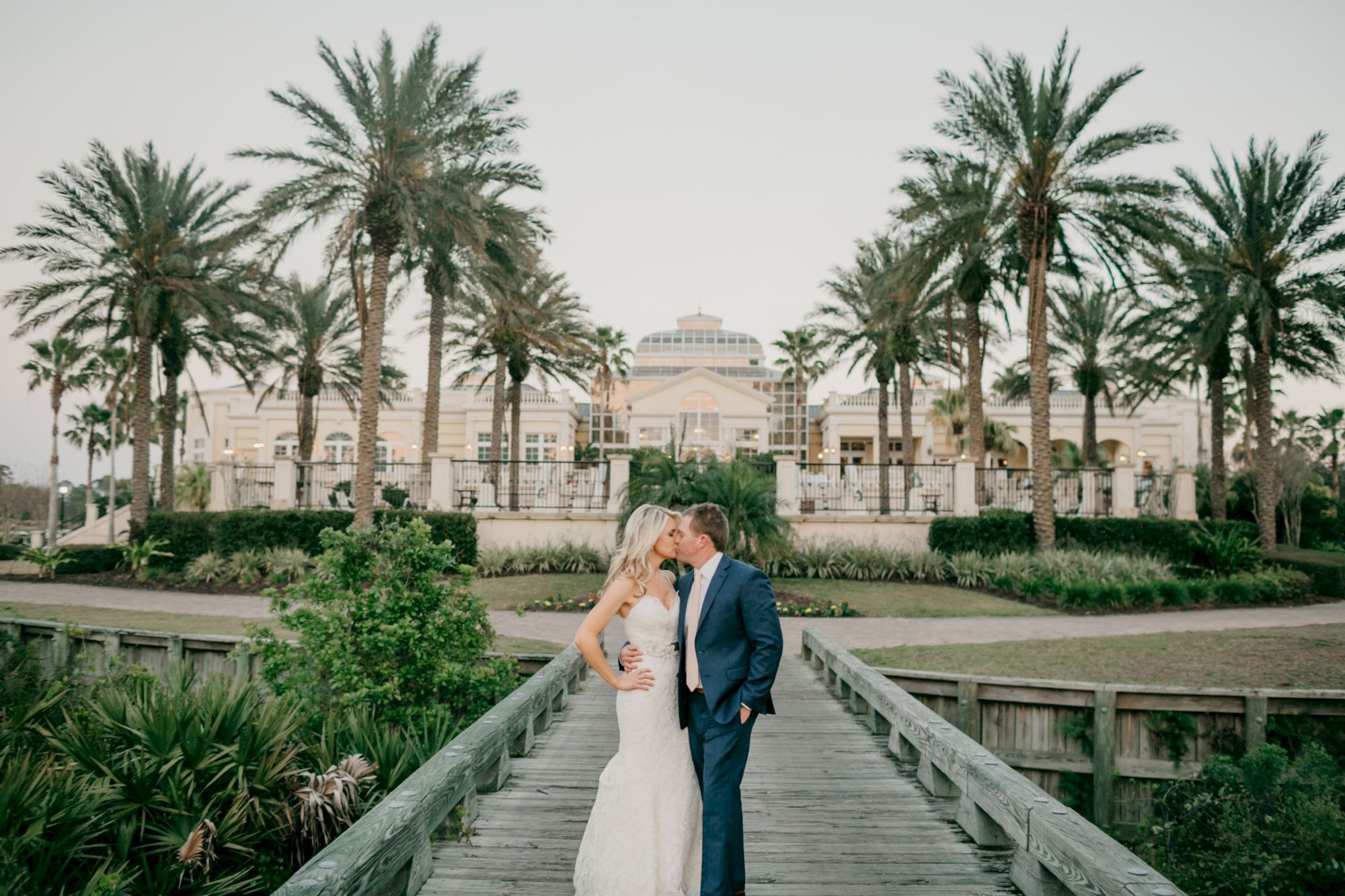 A couple is kissing on a wooden path surrounded by palm trees, with a grand building in the background.