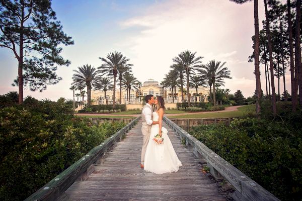 A couple in wedding attire kisses on a wooden bridge with a grand building and palm trees in the background.