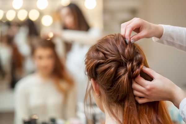 A hairstylist is braiding a woman's hair in a salon, with a mirror reflecting their image, creating a cozy and professional atmosphere.