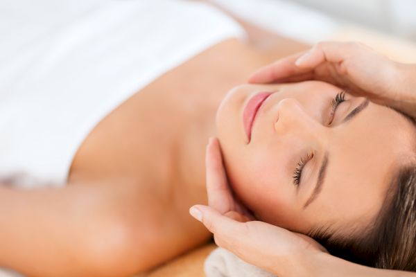 A person is receiving a facial massage, lying down with eyes closed, while hands gently cradle their face.