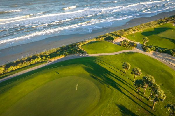 Aerial view of a coastal golf course featuring lush green fairways, palm trees, and ocean waves along a sandy shore.