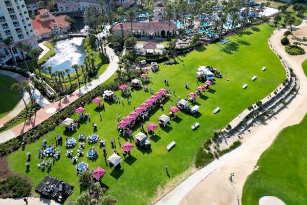 An outdoor event with tents, tables, and people on a lawn near a pond, surrounded by buildings and palm trees.