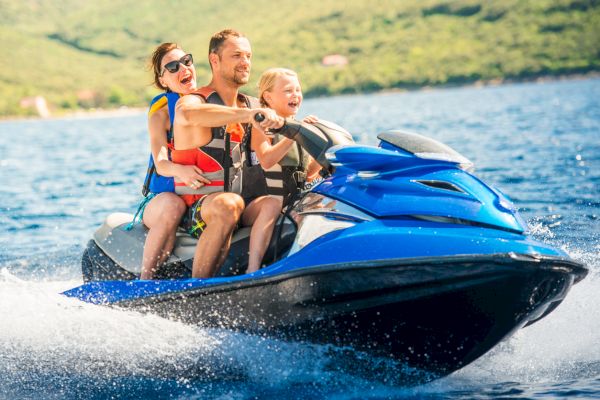 A family of three wearing life jackets rides a blue jet ski on a sunny day, enjoying the water and the scenery.