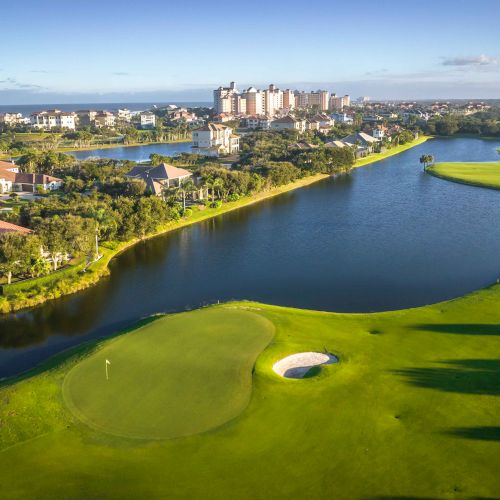 Aerial view of a golf course with a lake, green fairways, sand traps, surrounded by houses and buildings, under a clear blue sky.