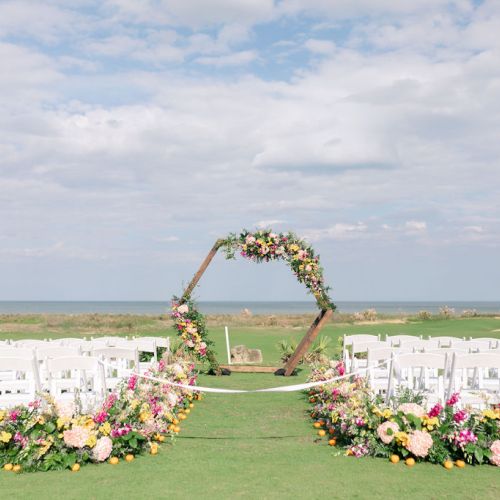 Outdoor wedding setup overlooking the sea, with a flower-decorated arch and rows of white chairs, on a grassy lawn.