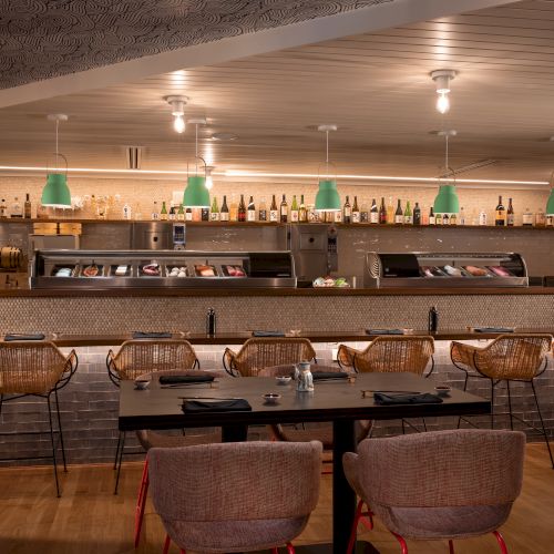 A sushi restaurant with a bar, green pendant lights, wicker chairs, bottles on shelves, sushi display, and wooden flooring.
