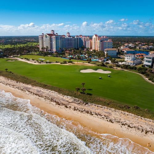 Beachfront golf course and a large resort with surrounding buildings under a clear blue sky.