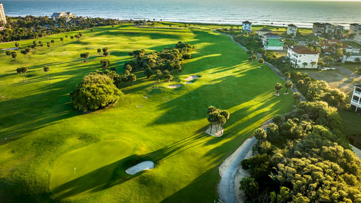 Aerial view of a coastal golf course with manicured greens, sand traps, trees, and ocean views, surrounded by residential buildings.