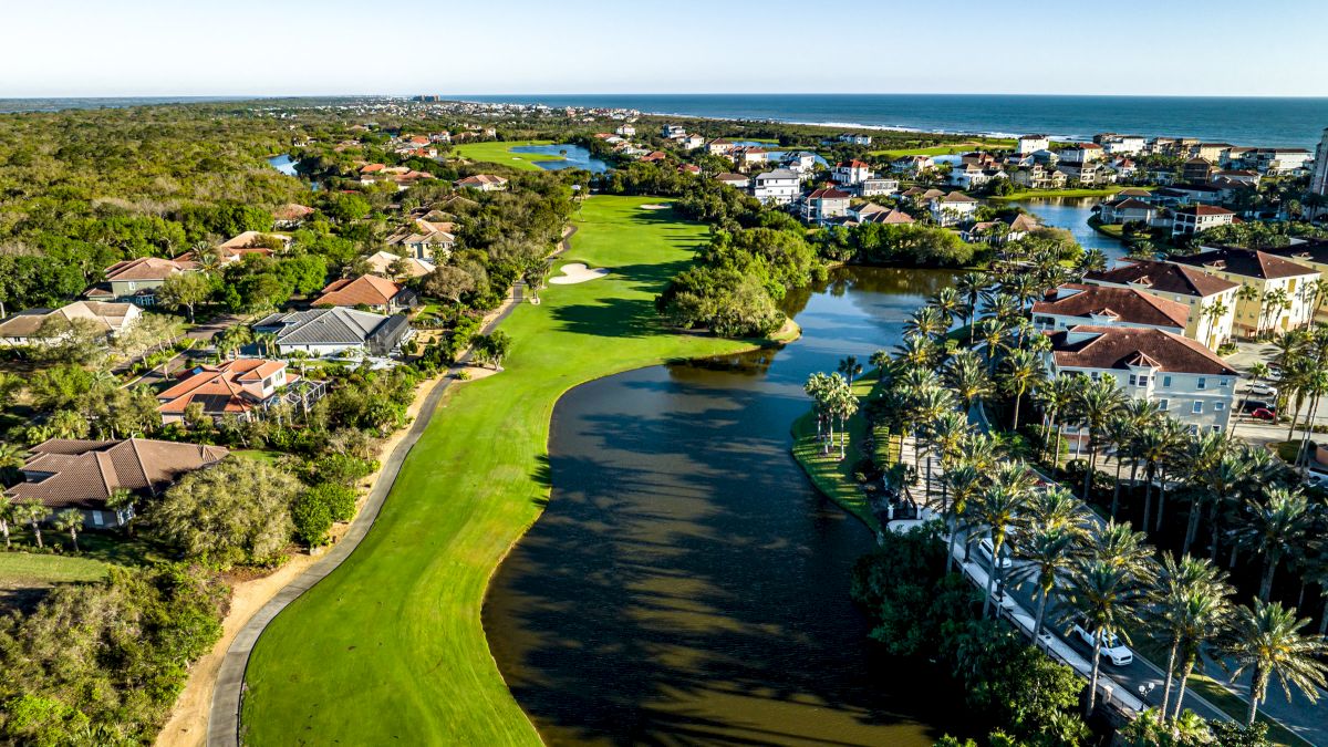 Aerial view of a coastal residential area with a golf course, lush greenery, palm trees, houses, and a glimpse of the ocean in the background.