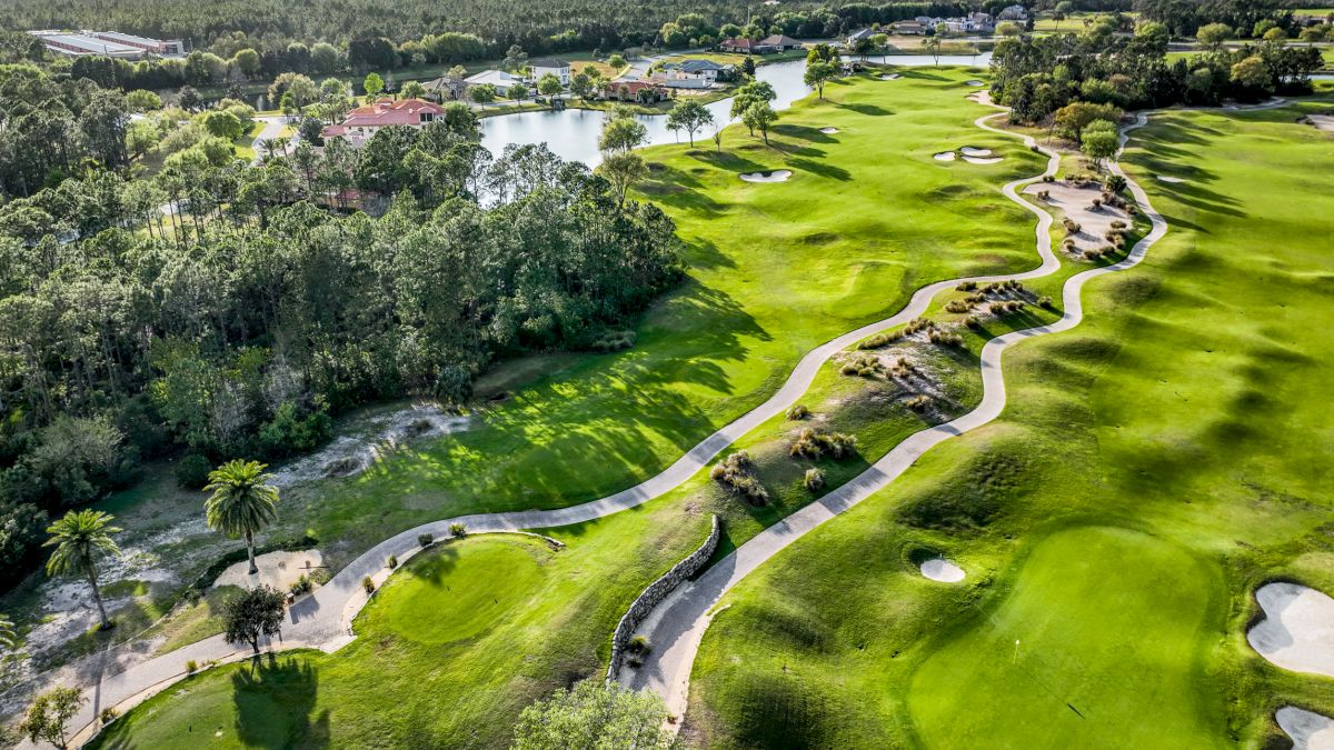 An aerial view of a scenic golf course with lush greens, winding paths, trees, and a lake, surrounded by forested areas.