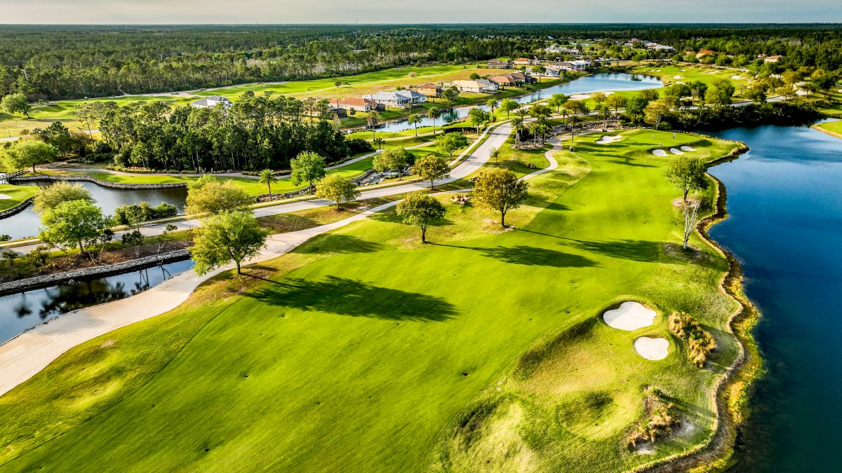 Aerial view of a golf course featuring lush green fairways, sand bunkers, water hazards, and a scenic landscape with trees and homes.