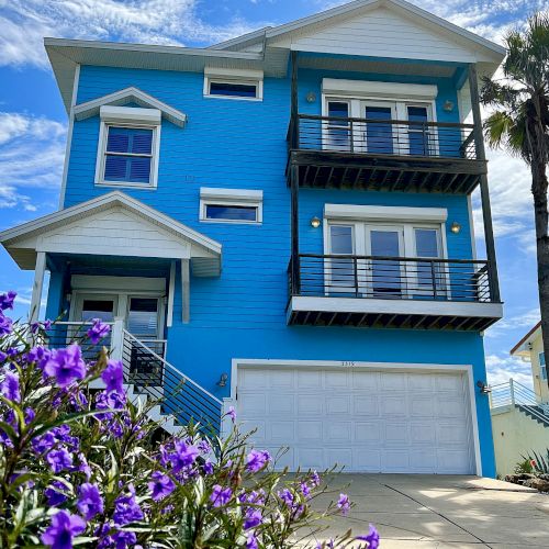 A vibrant blue house with balconies, surrounded by lush purple flowers and a tall palm tree under a clear blue sky.