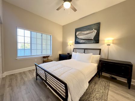 A bedroom with a bed, nightstands, a painting of a boat, a window with blinds, and a ceiling fan.
