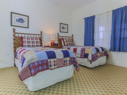 The image shows a bedroom with two twin beds, plaid bedspreads, blue curtains, fish artwork, a nightstand, and a red lamp.