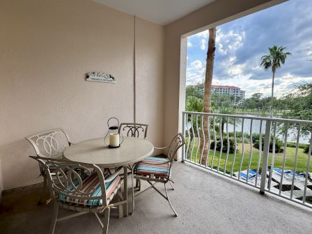 A balcony with a table, chairs, and a sign reading "Welcome to the Beach" overlooks a palm tree and greenery.