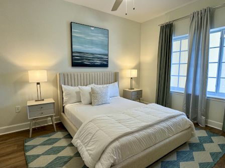 A cozy bedroom featuring a double bed, nightstands with lamps, artwork, curtains, and a patterned rug, in a softly lit space.