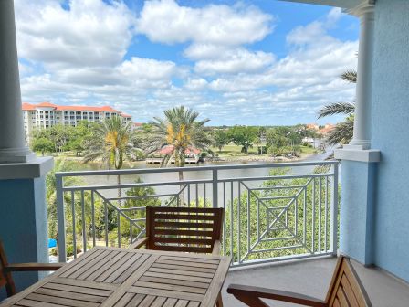 A balcony view with a wooden table and chairs, overlooking a river, palm trees, and buildings with red roofs under a partly cloudy sky.