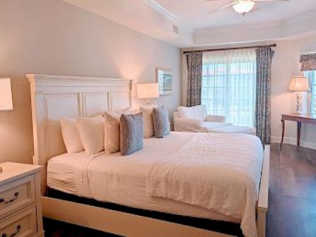 A cozy bedroom with a large bed, chairs, lamps, a desk, and patterned curtains. Natural light comes through the windows.
