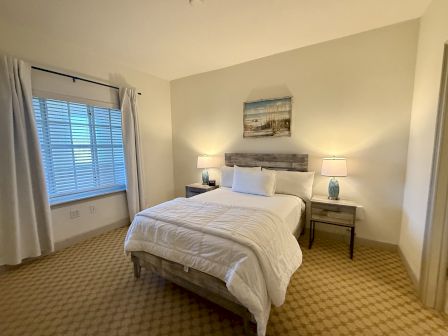 A tidy bedroom with a bed, two nightstands, lamps, a window with blinds and curtains, and a landscape painting above the bed.