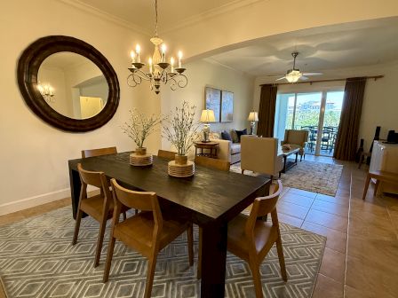 A dining room with a dark table, wooden chairs, and a round mirror; living area with couches and glass doors in the background.
