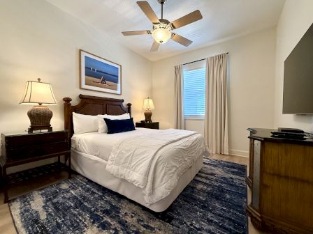 A cozy bedroom with a bed, two lamps, a ceiling fan, window with curtains, TV, and a beach-themed picture on the wall.