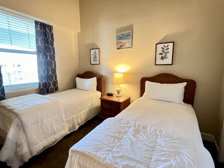 A cozy bedroom with two single beds, a bedside table with a lamp, decorative art, and a window with patterned curtains.