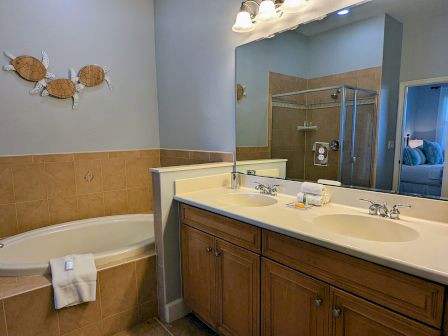 A bathroom with twin sinks, bathtub, glass shower, towels, turtle wall decor, and a reflection of the adjacent bedroom in the mirror.