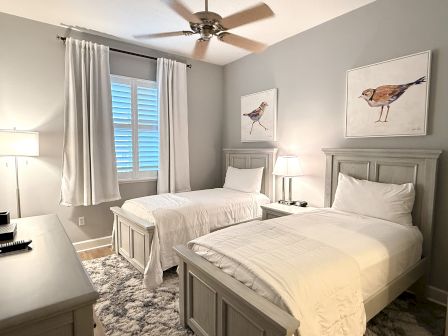 This image shows a bedroom with two single beds, white bed linens, bird artwork on the walls, a window with curtains, and a ceiling fan.