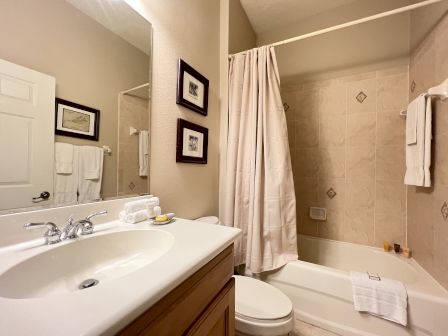 A bathroom with a sink, mirror, toilet, and a shower-tub combo with a curtain. Towels and framed pictures adorn the beige walls.