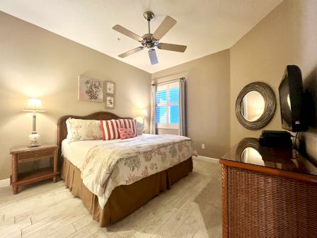 A cozy bedroom with a large bed, decorative pillows, side tables with lamps, wall art, a ceiling fan, a TV, and a mirror on the dresser.