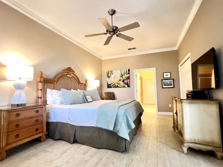 A cozy bedroom with a large bed, bedside tables with lamps, a ceiling fan, a dresser, and a TV. Art decorates the wall.