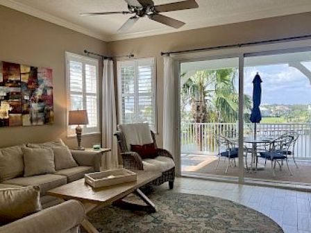 A cozy living room with a beige sofa, abstract art, and a rug next to a balcony with a table, chairs, and outdoor view.