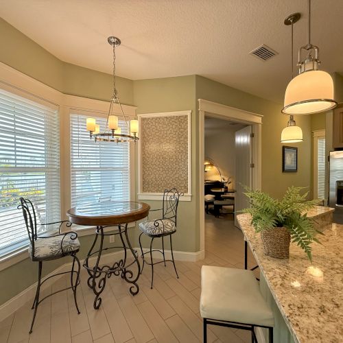 A cozy kitchen nook with a round table, cushioned chairs, and pendant lights. Granite counter has a potted fern; nearby stainless steel fridge.