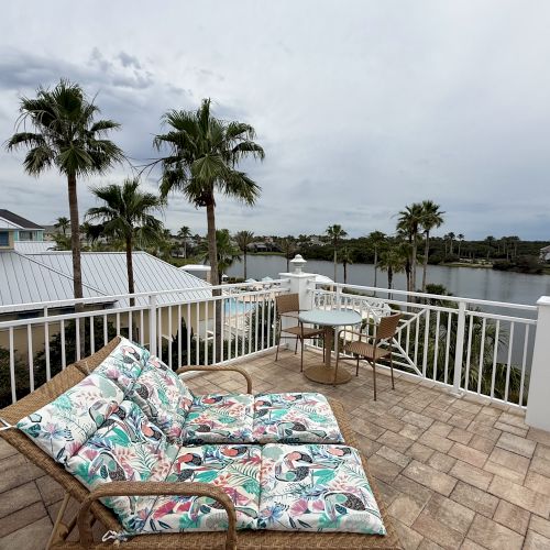 A patio with loungers and a table overlooks a scenic view with palm trees and a body of water, under a cloudy sky.