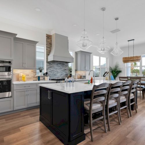A modern kitchen with gray cabinets, a large island with barstools, pendant lights, and dining area with a table and chairs in the background.