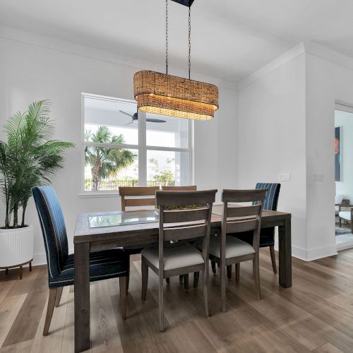 A modern dining room with a wooden table, six chairs, a hanging light fixture, potted plant, and large windows letting in natural light.