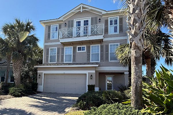 A two-story house with a garage, surrounded by palm trees and landscaped greenery, under a clear blue sky.