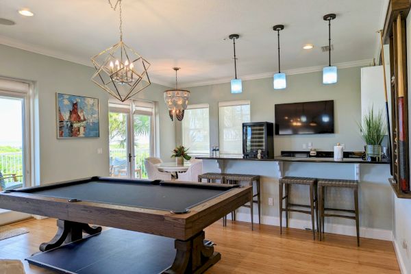 A stylish room features a pool table, a bar with stools, pendant lights, wall art, a TV, and a round dining table near large windows.