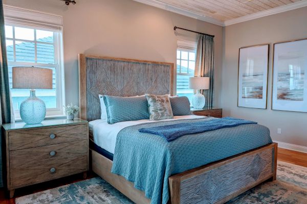 A cozy bedroom features a wooden bed with blue bedding, two nightstands with lamps, large windows, and abstract art on the walls.